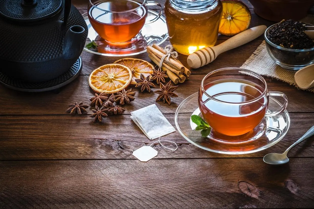 Xícaras de chá com canela, anis-estrelado, fatias de laranja seca, mel e sachês sobre uma mesa de madeira.