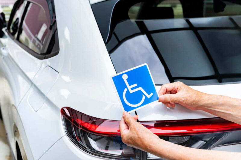 Pessoa idosa com as mãos visivelmente enrugadas segura um adesivo azul com o símbolo internacional de acessibilidade (cadeira de rodas) próximo à traseira de um carro branco moderno. A cena é iluminada por luz natural. O foco está na aplicação do adesivo próximo à lanterna traseira do veículo, sugerindo o preparo para sinalização de um automóvel destinado a pessoa com deficiência (PCD). O fundo mostra uma área coberta com estrutura metálica e vidro.
