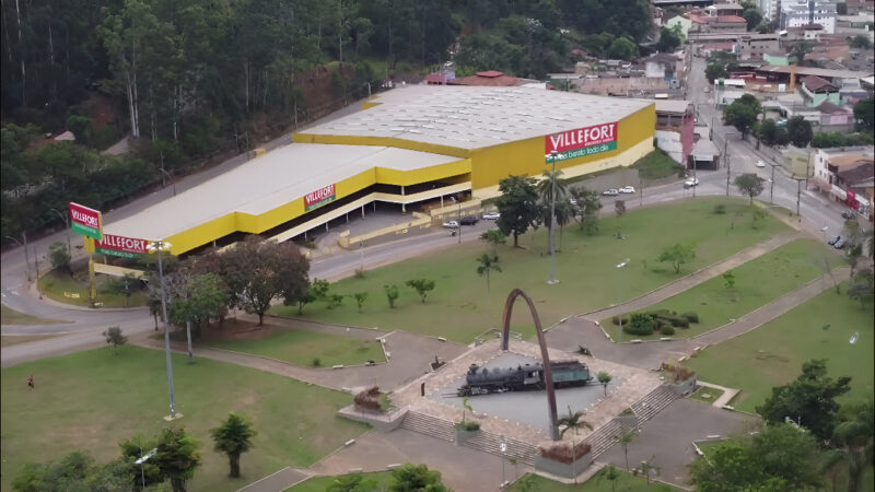 Imagem aérea mostrando a fachada do Villefort Atacadista em Itabira, Minas Gerais. O prédio é grande, com pintura amarela e letreiros vermelhos. Em primeiro plano, aparece a Praça do Centenário, com um monumento em forma de arco e esculturas, rodeada por áreas verdes e vias de acesso.