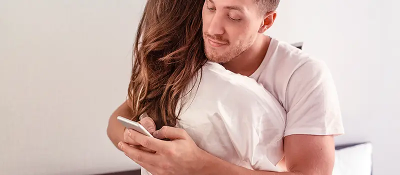 Homem abraça uma mulher enquanto olha para o celular com expressão suspeita, escondendo a tela dela.


