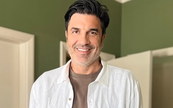 Edu Guedes sentado, sorrindo para a câmera, com boné azul e camiseta azul clara com estampa amarela.