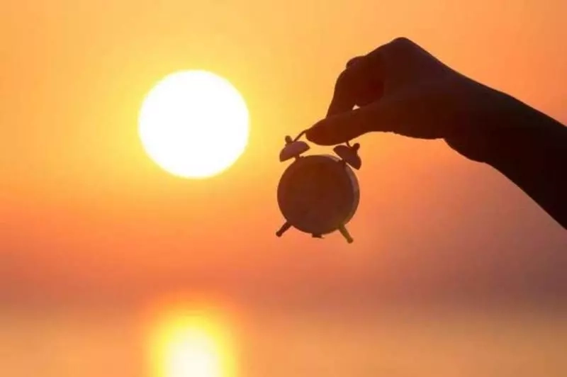 Relógio analógico antigo em primeiro plano, com sol forte ao fundo em meio à vegetação.

