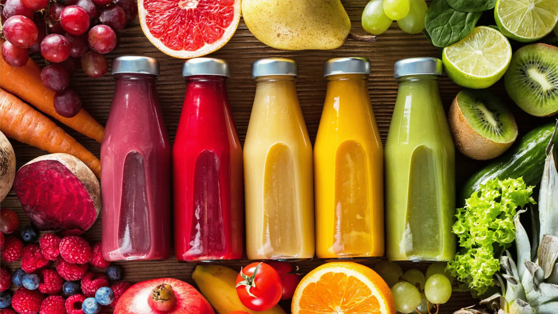 Cinco garrafas de sucos naturais de diferentes cores cercadas por frutas frescas e vegetais.
