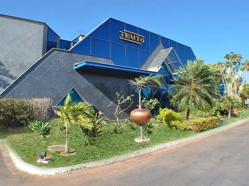 Entrada principal do Laboratório Teuto, com arquitetura moderna, jardim arborizado e o nome da empresa em destaque.